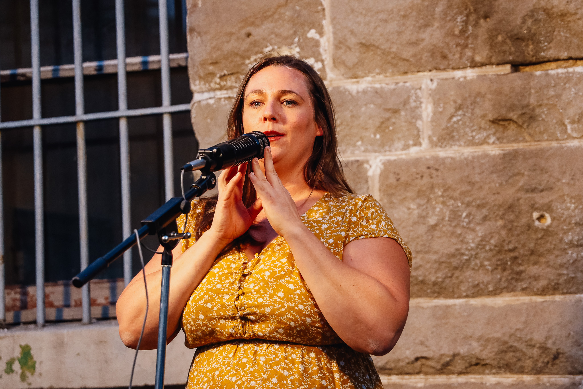 A woman in a yellow dress at a microphone in front of a stone wall and window with bars